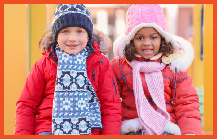 Photo of two children in orange-red winter jackets and scarves smiling at the camera.