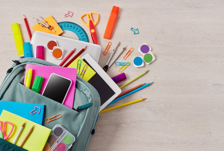 Photo of school supplies spilling out of a backpack lying on a pale wooden desk top.