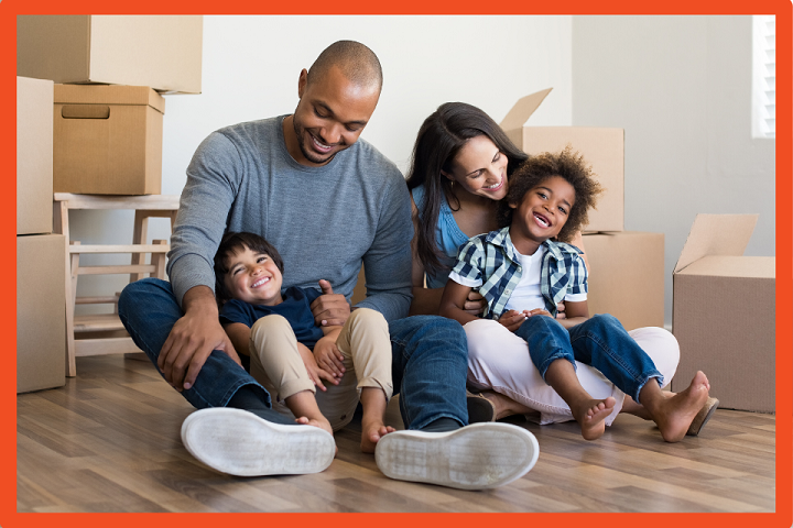 Photo of a family sitting together on the floor, smiling, with boxes piled behind them